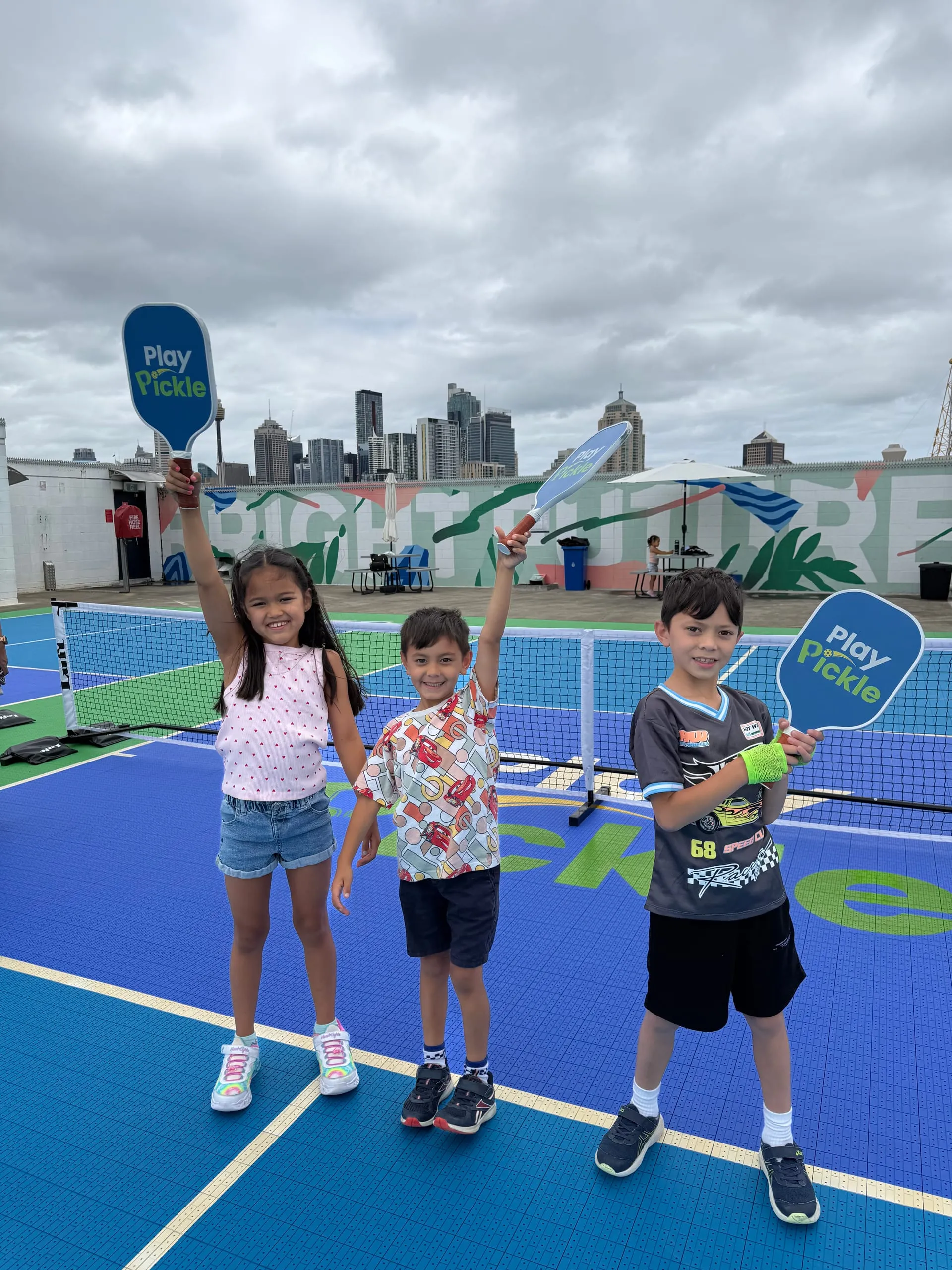 Three young players holding Play Pickle paddles above their heads on a rooftop court, with the Sydney skyline behind them