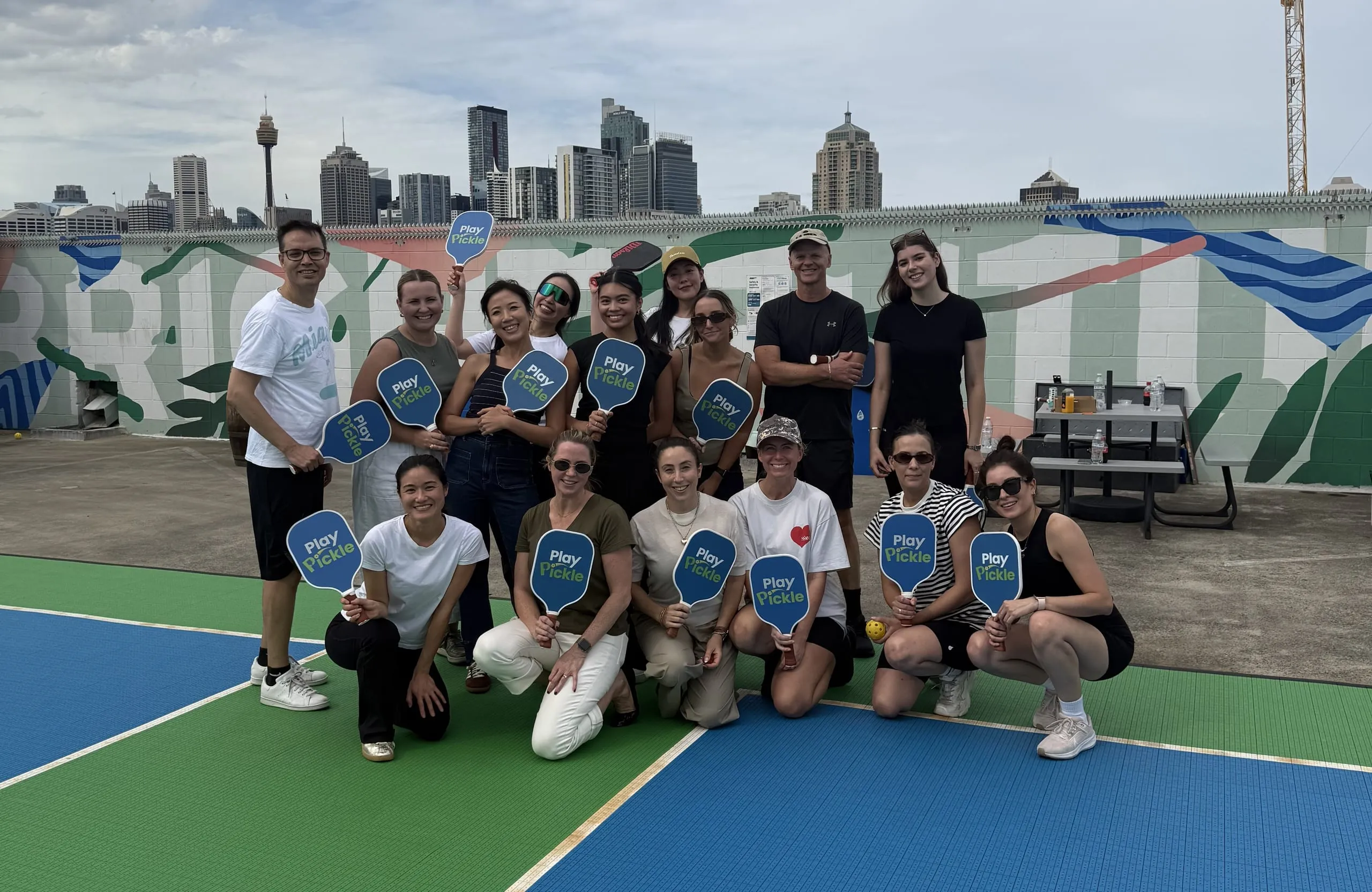 Play Pickle players on a rooftop court holding branded paddles, with the Sydney city skyline behind them