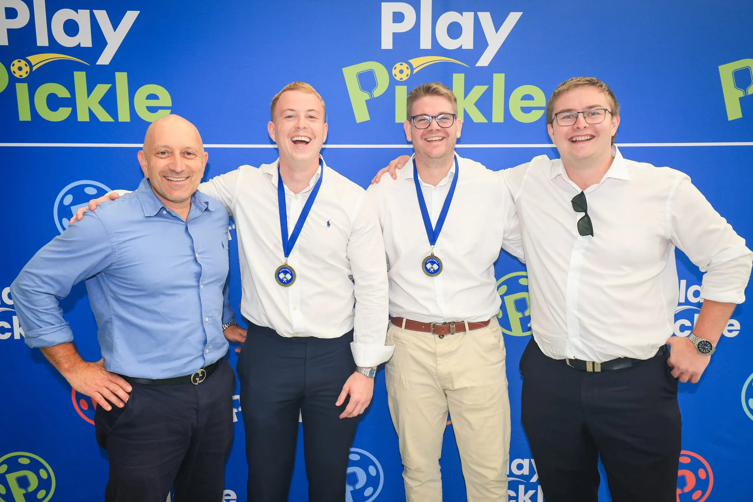 Four colleagues wearing medals and smiling in front of a Play Pickle branded backdrop at a corporate event